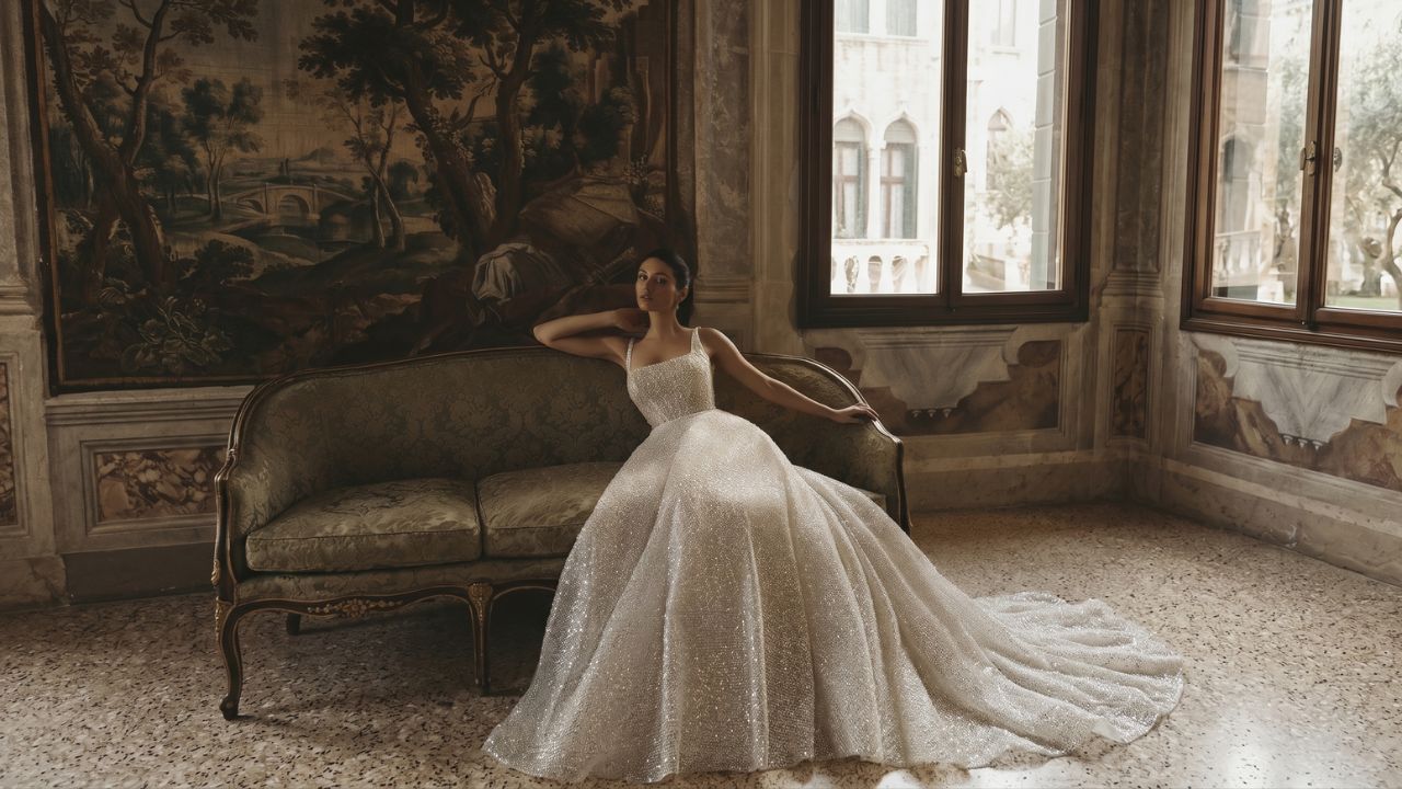Bride wearing a sparkling white ball gown wedding dress while seated on an antique sofa in an elegant historic room.